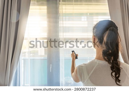 Woman using a remote control to adjust exterior window blinds in a modern apartment. Smart home comfort and energy efficient living concept