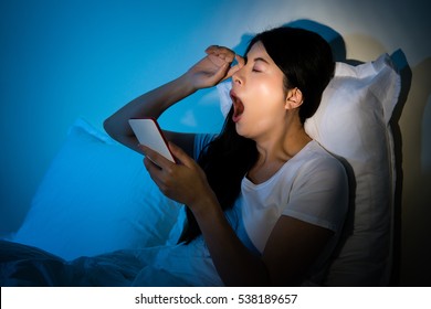 Woman Using Smarphone Working Rubbing Her Eyes And Yawn At Late Night Sitting On Bed. Mobile Addict Concept. Mixed Race Asian Chinese Model