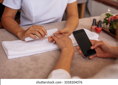 Woman Using Smart Phone While Receiving Manicure In Nail Salon. Female Client With Mobile Phone Getting Manicure And Nail Care Procedure By Beautician.