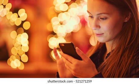 Woman Using Smartphone on European Christmas Market. 4K. Girl Enjoying Winter Holiday Season, Using phone, social networking, communicating, using app. Blurred Christmas Lights on background, dusk.