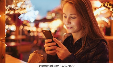 Woman Using Smartphone on European Christmas Market. 4K. Girl Enjoying Winter Holiday Season, Using phone, social networking, communicating, using app. Blurred Christmas Lights on background, dusk.