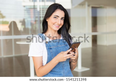 Woman using smartphone and smiling at camera. Beautiful cheerful young brunette woman using mobile phone and looking at camera outdoor. Technology concept