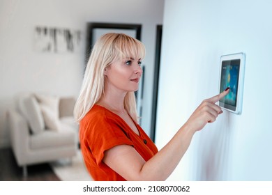 Woman Using Tablet Panel Application For Control Smart Home Living. Blonde Female Touch Wireless Device Controller For Home Automation On The Wall