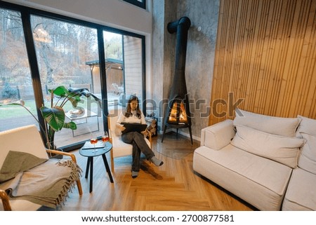 Woman using tablet while relaxing in modern living room with fireplace and large windows