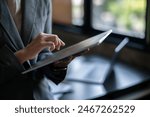 A woman is using a tablet while sitting at a desk. The tablet is placed on a table next to a laptop. The woman is wearing a suit and she is focused on her work
