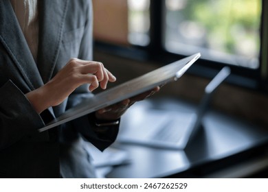 A woman is using a tablet while sitting at a desk. The tablet is placed on a table next to a laptop. The woman is wearing a suit and she is focused on her work - Powered by Shutterstock