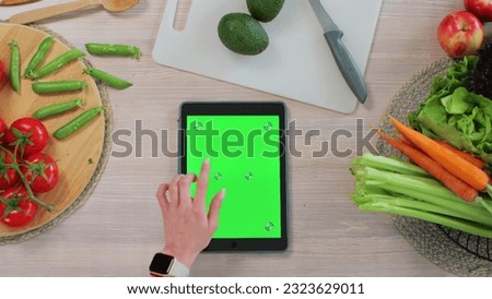 Woman using a tabletPC with a green screen at the kitchen table with fresh vegetables and fruits. Application for cooking. Healthy food concept.