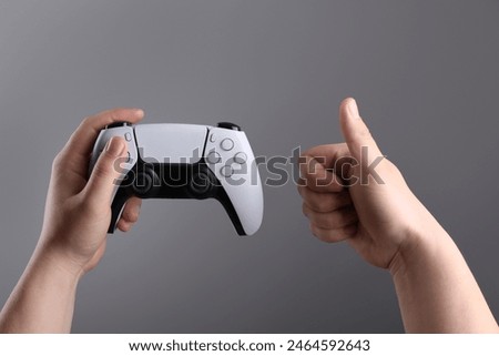 Woman using wireless game controller and showing thumbs up on grey background, closeup