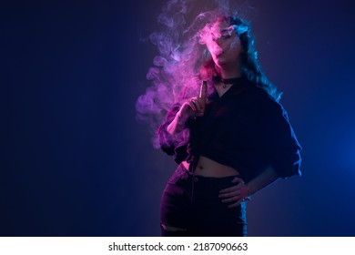 Woman with VAPE. Girl smokes VAPE and lets out smoke. Girl vaper on dark neon background. Woman with e-cigarette looks at smoke. Vaping concept with place for text. Vaper lady enjoys smoking