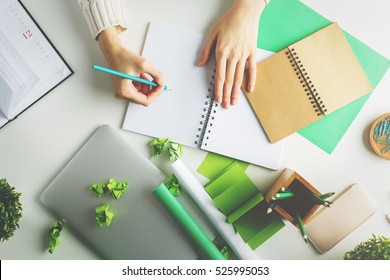 Woman With Various Creative Green Items On Desktop Writing In Spiral Notepad. Top View