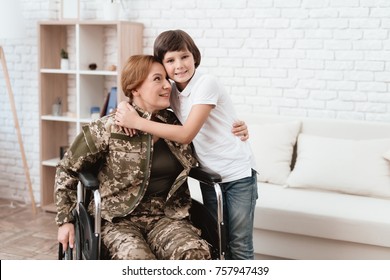 Woman Veteran In Wheelchair Returned Home. The Son Is Happy To See His Mother After Returning From The Army. Son Hugs Mom In Wheelchair.