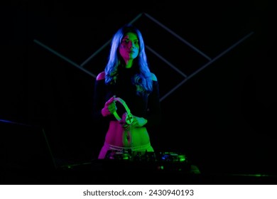 A woman with vibrant blue hair is DJing on a turntable. Holding headphones and looking at camera. Sidelit with colorful neon lights