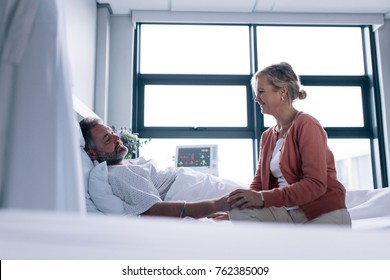 Woman visiting husband in hospital. Female talking with male patient lying in hospital bed.