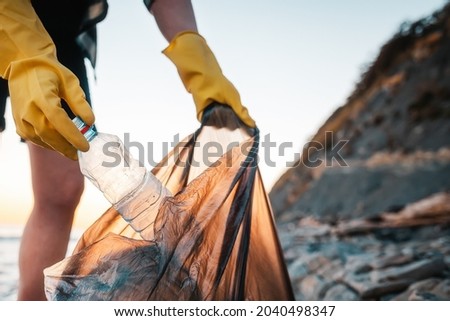 A woman volunteer puts a plastic bottle in a polyethylene bag. Close up of hands. In the background wild beach and ocean. The concept of environmental conservation and clean up of coast.