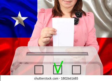 Woman Voter In A Smart Pink Jacket Lowers The Ballot In A Transparent Ballot Box Against The Background Of The National Flag Of The Country Chile, Concept Of State Elections, Referendum