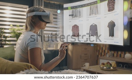 Woman in VR headset chooses clothes in online store chilling at home. 3D futuristic hologram shows widgets in user menu and clothing store website interface. VFX animation. Concept of online shopping.