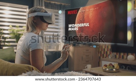 Woman in VR headset chooses movie or TV show to watch at home. 3D futuristic hologram shows interface of streaming service application and widgets in user menu. VFX animation. Concept of lifestyle.