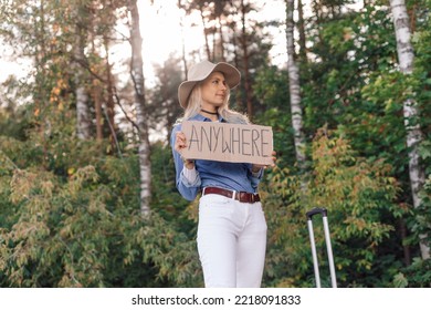 Woman Wait Passing Car In Forest Standing With Suitcase And Cardboard Poster On Roadside Of Highway. Blonde Lady In Hat Escape From City To Go Anywhere. Travelling, Auto Stop, Hitchhiking, Vacations.