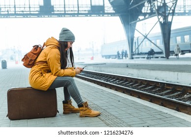 Woman Wait For Train On Railway Station. Travel Concept. Surfing Internet On Phone To Kill Time