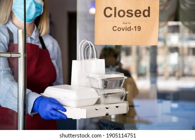 Woman Waiter In Protective Medical Mask And Gloves Work With Takeaway Orders. Waiter Giving Takeout Meal While Lockdown, Coronavirus Shutdown. Food For Delivery Against Sign Closed Due To Covid 19