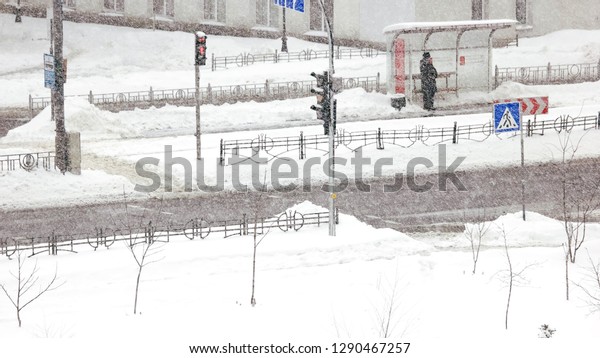 Woman is waiting for the bus during\
snow storm. City road during a heavy blizzard in winter. Bad winter\
weather conditions. Urban scene of city life in\
winter.