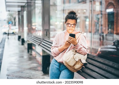 Woman waiting for public transport on bus stop looking to the smartphone, using application for searching traffic schedule. 