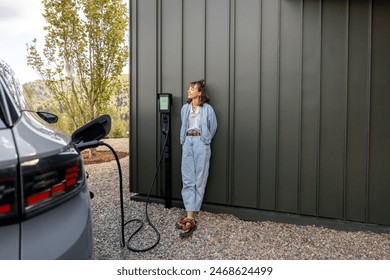 Woman waits for electric car to be charged standing near a charging station leaning to a house wall. Concept of modern lifestyle and smart technologies