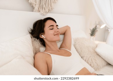 woman wake up in morning in boho style bedroom, sitting on bed, listening to music on earpods, relaxing