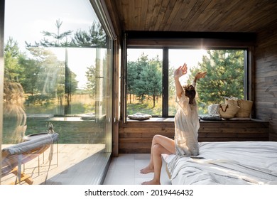 Woman wakes up in a country house or hotel with panoramic windows in pine forest raised her hands yawning. Good morning and recreation on nature concept