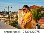 Woman is walking alone on sunny promenade with palm trees and flowers. Female tourist strolls along marina at sunset in coastal town