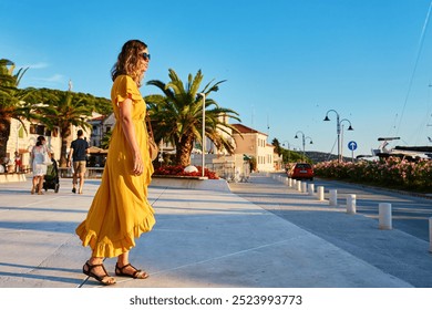 Woman is walking alone on sunny promenade with palm trees and flowers. Female tourist strolls along marina at sunset in coastal town - Powered by Shutterstock