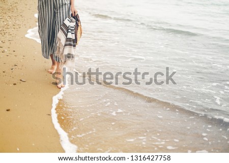 Woman walking barefoot on beach, close up view of legs and waves. Young girl relaxing on sandy beach near sea, walking with bag in hand. Summer vacation concept