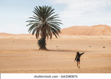 Woman Walking In Beautiful Desert With Sand Dunes And One Lonely Palm. View From Behind. Freedom And Travel Concept.