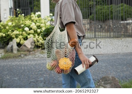 Woman walking and carrying white mesh string knitted shopping bag with groceries and reuseble metal cup. Eco friendly, reusable shopping bag. Zero waste and plastic free concept.