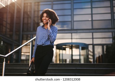 Woman Walking Down The Steps Outside Office Building And Talking On Mobile Phone. Businesswoman Talking On Cell Phone With Office Building In Background.