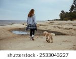 Woman walking her dog along the shore, wrapped in warm autumn clothing. The serene beach setting creates a peaceful outdoor mood, perfect for themes of nature and pet care.