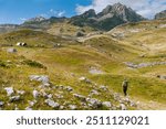 Woman walking in mountainous valley with huts and view of mountain peaks. Female tourist traveling with backpack and trekking poles, view from back. Healthy lifestyle and active people concept.