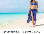 Woman walking on beach in blue fashion beachwear bathing suit and sarong pareo sun skirt relaxing in luxury Caribbean vacation holidays. Summer or winter getaway destination.