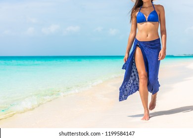 Woman walking on beach in blue fashion beachwear bathing suit and sarong pareo sun skirt relaxing in luxury Caribbean vacation holidays. Summer or winter getaway destination.
