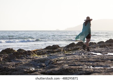 woman walking on rocky beach at sunset with sun hat, heels, and large flowing sweater/ woman on rocky beach/ woman on rocky beach at sunset