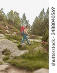 A woman is walking on a rocky path with a backpack and a walking stick. The path is surrounded by trees and bushes, and there are flowers growing along the way. The woman is enjoying her hike