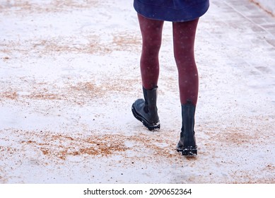 Woman Walking On Snowy Sidewalk Spreading With Sand, Prevention Of Pedestrian Injury. Back View On Female Feet In Winter Black Boots Walking On Snowy And Icy Walkway. Slippery Road Sprinkled With Sand