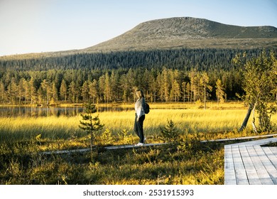 Woman walking on a wooden path beside a serene lake in the Idre wetlands, with the majestic Mount Städjan rising in the background during the golden hour of summer in Dalarna Sweden - Powered by Shutterstock