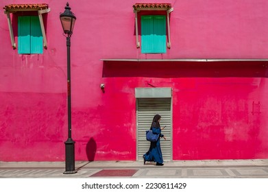 Woman walking past bright pink wall wearing a hijab carrying a blue tote bag and looking at a cell phone - Powered by Shutterstock