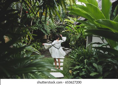 Woman Walking In Tropical Garden In White Dress. Brunette With Long Black Hair. Fashion Tunic, Barefoot, Enjoying Nature