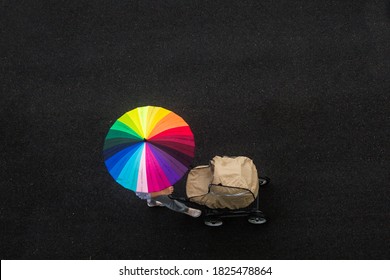 Woman Walks Along A Black Road Under An Umbrella Painted In The Form Of A Rainbow, Pushing A Stroller With A Child 