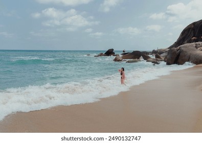 A woman walks along a sandy beach with white foamy waves crashing on the shore. In the background, rocky cliffs rise up against a clear blue sky with fluffy white clouds. - Powered by Shutterstock