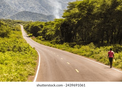 A woman walks down a long road in the woods. The road is empty and the trees are lush and green, Kenya. - Powered by Shutterstock