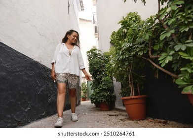 A woman walks joyfully in a narrow alley with lush plants. She wears casual summer clothes and exudes happiness in this cozy urban scene. - Powered by Shutterstock