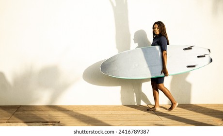 A woman walks near a wall with a surfboard dressed in a wetsuit. Concept of selling surfboards, advertising surfing equipment. Copy space for water sports ads. - Powered by Shutterstock